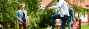 Three men play with a soccer ball on a grassy lawn, with a house and trees in the background.