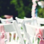 White chairs adorned with pink and blue ribbons arranged for an outdoor event with green foliage in the background.