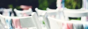 White chairs adorned with pink and blue ribbons arranged for an outdoor event with green foliage in the background.