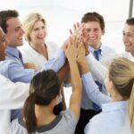 A group of eight people in business attire stand in a circle, smiling and performing a group high-five in an office setting.