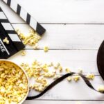 Film reel, clapperboard, and spilled popcorn on a white wooden surface, creating a movie-themed setting.