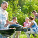 Older man grilling food outdoors, while four people sit at a table in the background enjoying a meal in a garden setting.