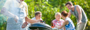 Older man grilling food outdoors, while four people sit at a table in the background enjoying a meal in a garden setting.