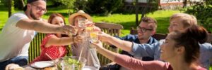 A group of six people sitting at an outdoor table, toasting with drinks, surrounded by greenery and a small house in the background.