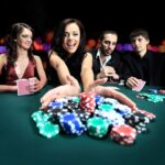 Four people at a poker table, one woman pulling in a pile of colorful poker chips, with a blurred bokeh background.