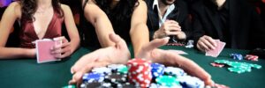 Four people at a poker table, one woman pulling in a pile of colorful poker chips, with a blurred bokeh background.