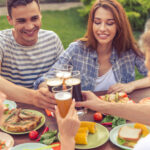 Four people enjoying a meal outdoors, clinking glasses of beer over a table filled with plates of food.