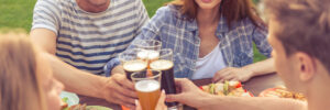 Four people enjoying a meal outdoors, clinking glasses of beer over a table filled with plates of food.