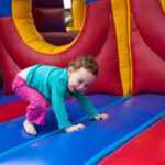 Child playing inside a colorful inflatable bounce house, crawling with a smile, wearing a blue shirt and pink pants.