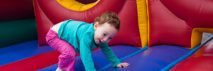 Child playing inside a colorful inflatable bounce house, crawling with a smile, wearing a blue shirt and pink pants.