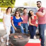 Group of five friends enjoying a roadside barbecue beside a pickup truck, holding drinks and a football.