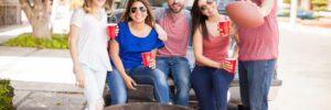 Group of five friends enjoying a roadside barbecue beside a pickup truck, holding drinks and a football.