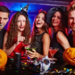 Five people in Halloween costumes smiling at a party with glowing jack-o'-lanterns and drinks on the table.