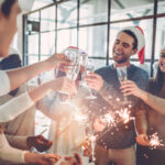 A group of people wearing Santa hats raise glasses and hold sparklers while celebrating indoors.