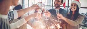 A group of people wearing Santa hats raise glasses and hold sparklers while celebrating indoors.