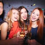 A group of five people smiling and holding glasses of champagne at a party, with colorful confetti in the background.