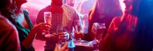 Group of people socializing and holding drinks, surrounded by colorful lights, in a lively party setting.