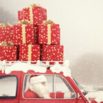 Santa driving a red car with snow and stacked red gift boxes tied to the roof, set against a snowy background.