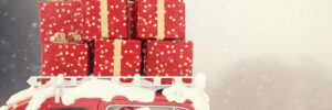 Santa driving a red car with snow and stacked red gift boxes tied to the roof, set against a snowy background.