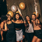 Five people smiling and holding sparklers while standing in front of gold and silver balloons.