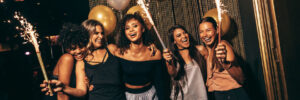 Five people smiling and holding sparklers while standing in front of gold and silver balloons.