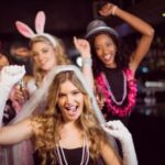 Four women at a bachelorette party, with one wearing a veil and gloves, and the others in festive accessories, holding drinks.