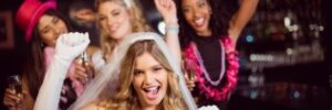 Four women at a bachelorette party, with one wearing a veil and gloves, and the others in festive accessories, holding drinks.