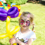 A child with butterfly face paint holds a yellow and purple balloon flower in a grassy area.