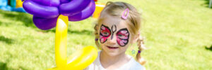 A child with butterfly face paint holds a yellow and purple balloon flower in a grassy area.