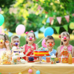 Children at a table wearing clown noses with balloons and party decorations in the background.