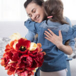 Woman and child embracing on a sofa, smiling, with a bouquet of red and yellow flowers in the foreground.