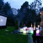 Outdoor movie setup with inflatable screen, seating, and patio heaters in a garden setting near a house at dusk.