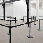 An empty queue system with black barriers forms a zigzag pattern in a large, brightly lit room with white walls.