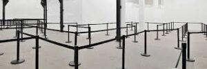 An empty queue system with black barriers forms a zigzag pattern in a large, brightly lit room with white walls.