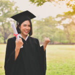 A graduate in a cap and gown holds a diploma and gestures a fist in celebration, standing outdoors in a grassy area with trees.