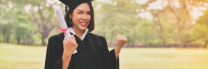 A graduate in a cap and gown holds a diploma and gestures a fist in celebration, standing outdoors in a grassy area with trees.