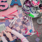 Hands organizing patriotic crafts with ribbons, paper, and USA letters on a wooden table.