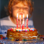 Child blowing out candles on a cake decorated with colorful candies, set on a glass stand.