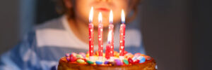 Child blowing out candles on a cake decorated with colorful candies, set on a glass stand.