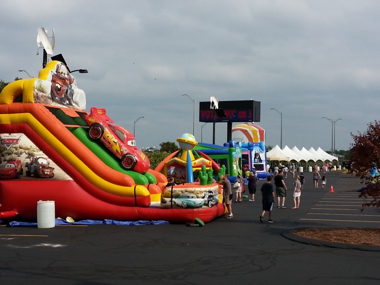 People walk through a parking lot with inflatable slides, tents, and overcast skies.