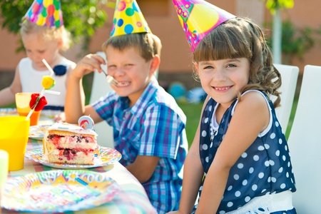 Children wearing party hats sit at a table outdoors with plates of cake, smiling and enjoying a birthday celebration.