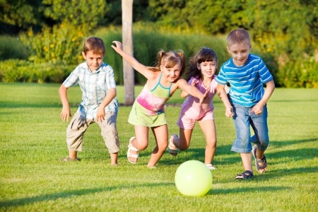 Four children run towards a green ball on a grassy field, surrounded by trees in the background.