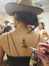 Person with a dragon tattoo on their back being sprayed; wearing a cowboy hat and tank top in a casual indoor setting.