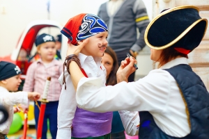 Children dressed as pirates, with one adult applying paint to a child's face, in a playful indoor setting.