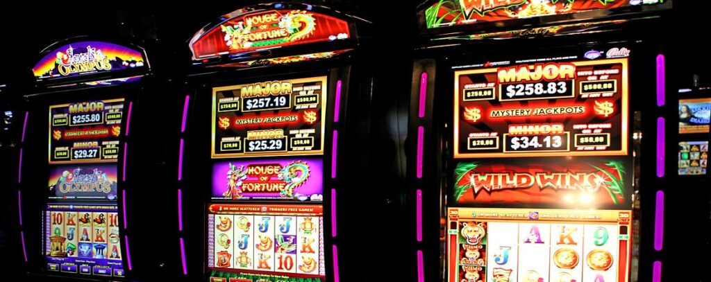 Three illuminated slot machines with colorful displays, showing various games and jackpot amounts in a casino setting.