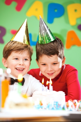 Two young boys wearing party hats smile excitedly at a lit birthday cake, with colorful decorations in the background.