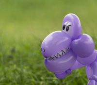 A purple balloon sculpture of a dinosaur with cartoonish eyes and teeth is set against a grassy background.