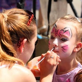 Child receiving face painting as a cat with a bow, done by a woman using a paintbrush, at an outdoor event.