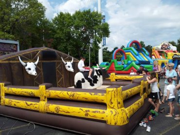 A person rides a mechanical bull at an outdoor amusement area with inflatable slides, surrounded by spectators.