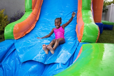 Child in a pink swimsuit sliding down a colorful inflatable water slide outdoors, smiling with arms raised.